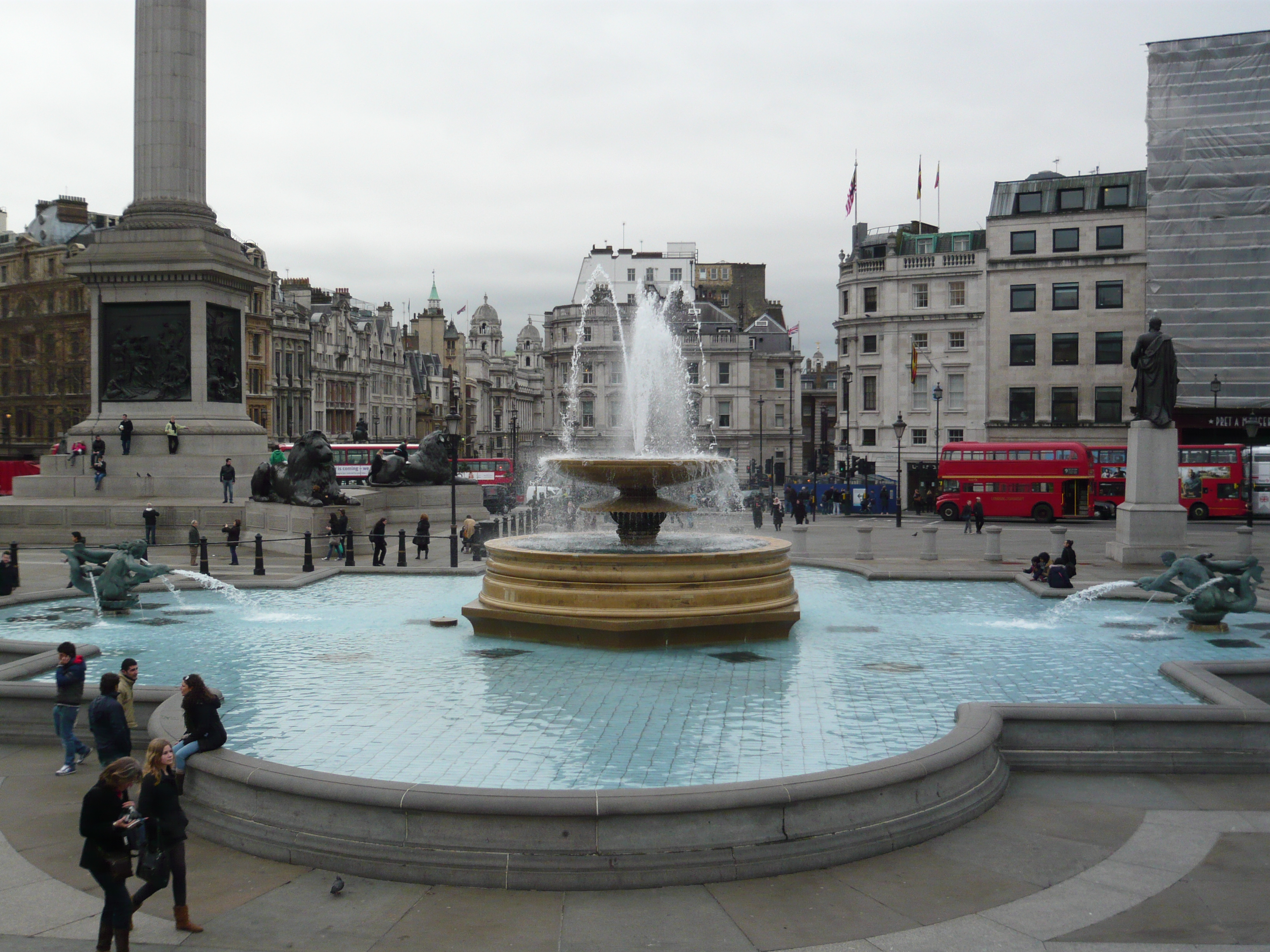 Trafalgar square fountain, red buss, people, grey day.